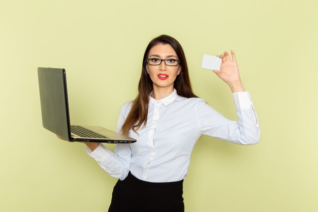 Vorderansicht des weiblichen Büroangestellten im weißen Hemd und im schwarzen Rock unter Verwendung ihres Laptops und des Haltens der Karte auf hellgrüner Wand