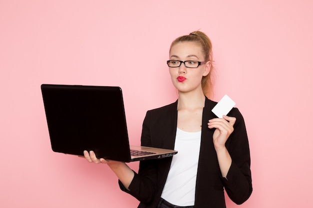 Vorderansicht des weiblichen büroangestellten in der schwarzen strengen jacke, die karte hält und laptop auf rosa wand verwendet