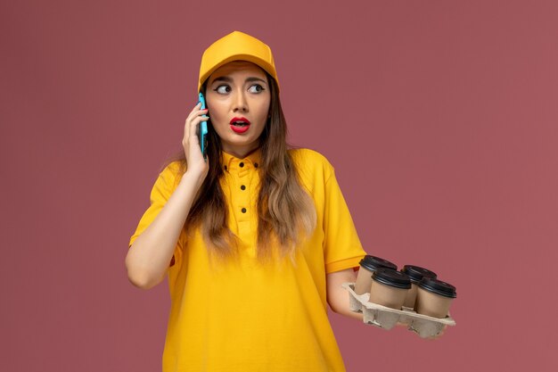 Vorderansicht des weiblichen Kuriers in der gelben Uniform und in der Kappe, die braune Kaffeetassen halten und am Telefon auf rosa Wand sprechen