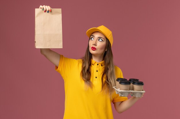 Vorderansicht des weiblichen Kuriers in der gelben Uniform und in der Kappe, die braune Kaffeetassen und Nahrungsmittelpaket auf der rosa Wand halten