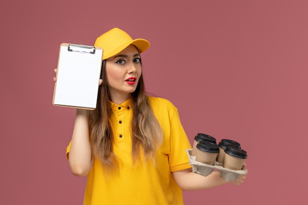 Vorderansicht des weiblichen Kuriers in der gelben Uniform und in der Kappe, die braune Kaffeetassen und Notizblock an der rosa Wand halten