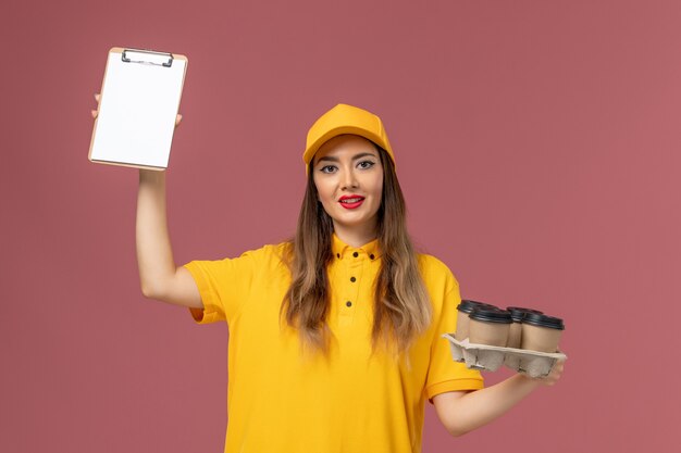 Vorderansicht des weiblichen Kuriers in der gelben Uniform und in der Kappe, die braune Kaffeetassen und Notizblock an der rosa Wand halten