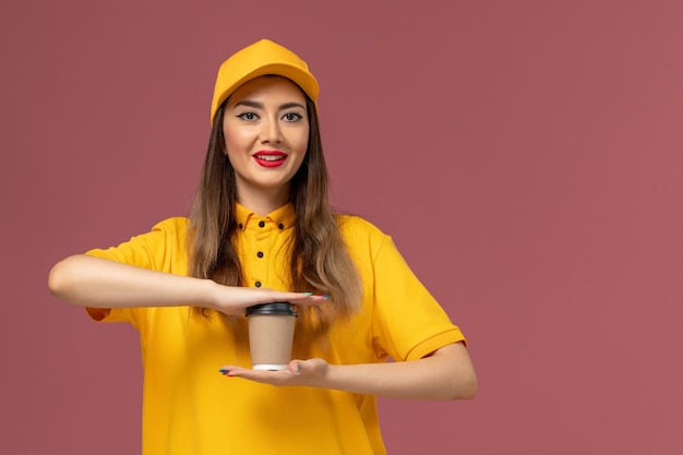 Vorderansicht des weiblichen Kuriers in der gelben Uniform und in der Kappe, die die Kaffeetasse der Lieferung an der rosa Wand hält