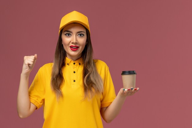 Vorderansicht des weiblichen Kuriers in der gelben Uniform und in der Kappe, die die Kaffeetasse der Lieferung an der rosa Wand hält