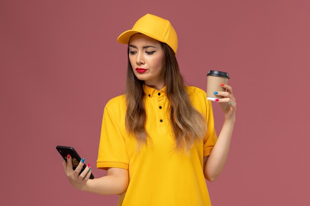 Vorderansicht des weiblichen Kuriers in der gelben Uniform und in der Kappe, die die Kaffeetasse der Lieferung hält und ein Telefon auf rosa Wand verwendet