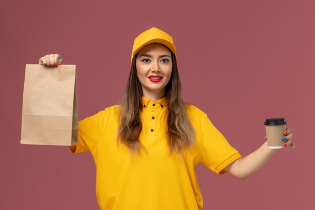 Vorderansicht des weiblichen Kuriers in der gelben Uniform und in der Kappe, die die Kaffeetasse und die Lebensmittelverpackung der Lieferung auf rosa Wand hält