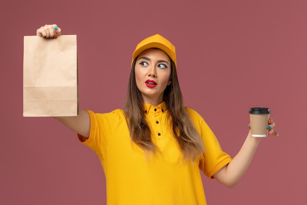 Vorderansicht des weiblichen Kuriers in der gelben Uniform und in der Kappe, die die Kaffeetasse und die Lebensmittelverpackung der Lieferung auf rosa Wand hält