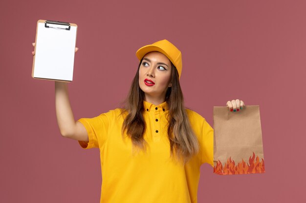 Vorderansicht des weiblichen Kuriers in der gelben Uniform und in der Kappe, die Lebensmittelpaket und Notizblock auf der rosa Wand hält