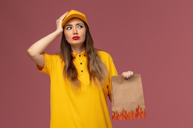 Vorderansicht des weiblichen Kuriers in der gelben Uniform und in der Kappe, die Lebensmittelverpackung hält und auf hellrosa Wand denkt