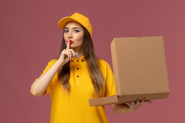 Vorderansicht des weiblichen Kuriers in der gelben Uniform und in der Kappe, die Nahrungsmittelbox auf der hellrosa Wand hält