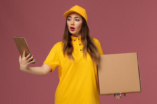 Vorderansicht des weiblichen Kuriers in der gelben Uniform und in der Kappe, die Nahrungsmittelbox und Notizblock auf rosa Wand hält