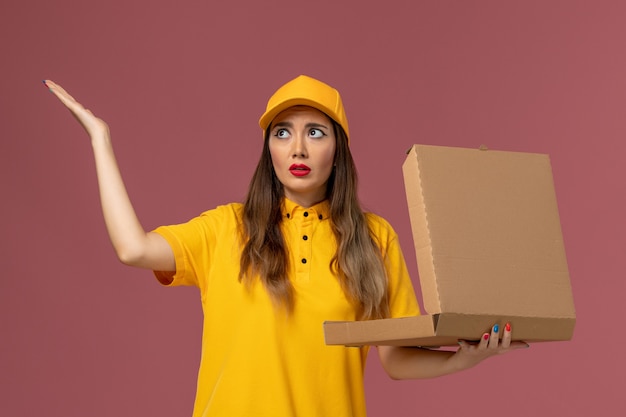 Kostenloses Foto vorderansicht des weiblichen kuriers in der gelben uniform und in der kappe, die offene nahrungsmittelbox auf der hellrosa wand hält