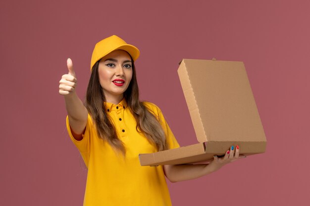 Vorderansicht des weiblichen Kuriers in der gelben Uniform und in der Kappe, die offene Nahrungsmittelbox auf der hellrosa Wand hält