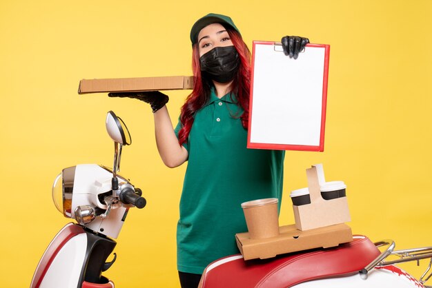 Vorderansicht des weiblichen Kuriers in der Maske mit dem Lieferkaffee und dem Essen an der gelben Wand