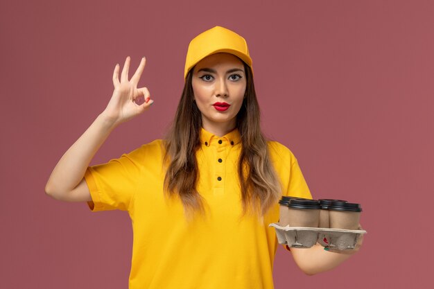 Vorderansicht des weiblichen Kuriers in gelber Uniform und Kappe, die braune Lieferkaffeetassen an der rosa Wand halten