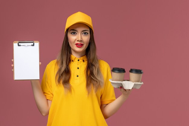 Vorderansicht des weiblichen Kuriers in gelber Uniform und Kappe, die Lieferkaffeetassen-Notizblock auf hellrosa Wand hält