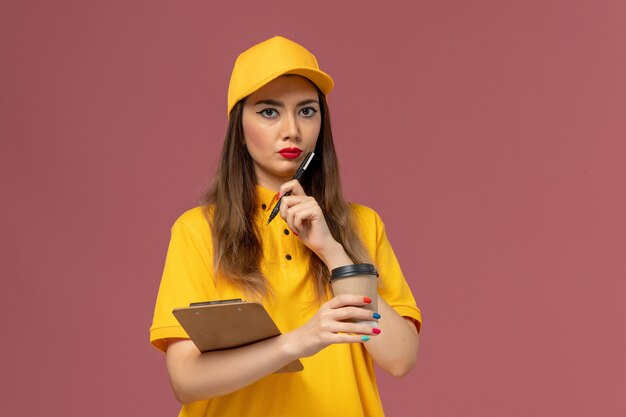 Vorderansicht des weiblichen Kuriers in gelber Uniform und Kappe, die Lieferung Kaffeetasse und Notizblock mit Stift auf der rosa Wand hält