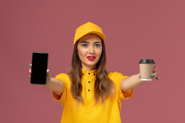 Vorderansicht des weiblichen Kuriers in gelber Uniform und Kappe, die Lieferung Kaffeetasse und Telefon auf rosa Wand hält