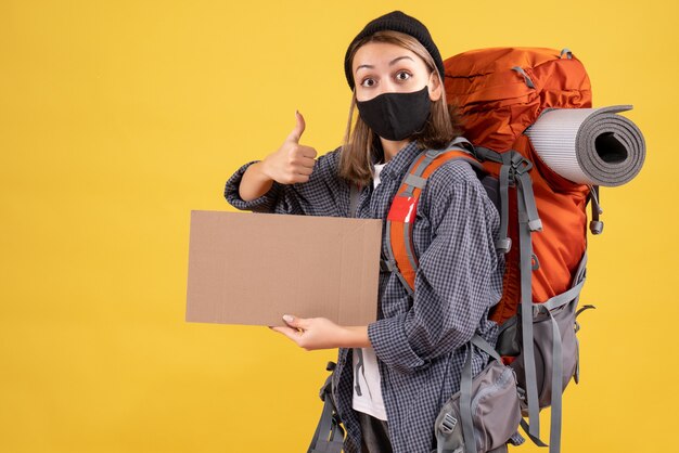 Vorderansicht des weiblichen Reisenden mit der schwarzen Maske und dem Rucksack, die Pappe halten, die Daumen hoch Zeichen machen