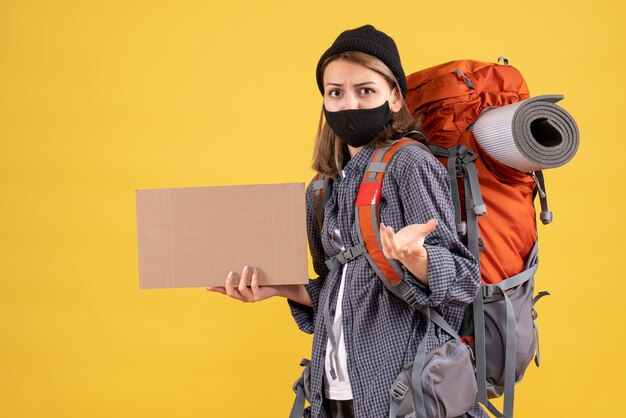 Vorderansicht des weiblichen Reisenden mit der schwarzen Maske und dem Rucksack, die Pappe halten