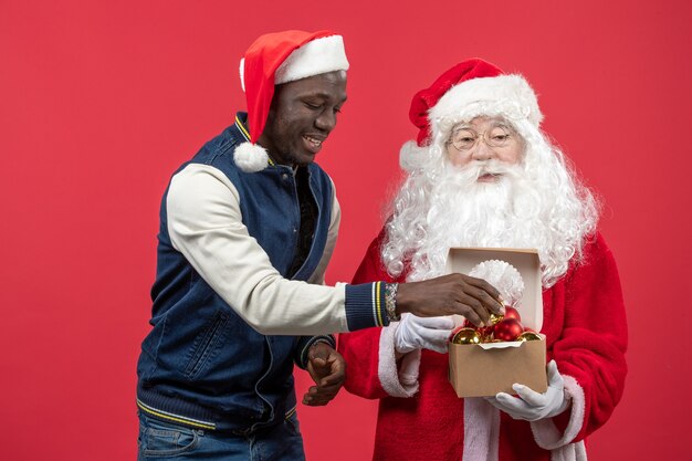 Vorderansicht des Weihnachtsmannes mit dem jungen jungen Mann, der Weihnachtsbaumspielzeug an der roten Wand hält