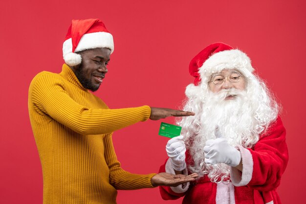Vorderansicht des Weihnachtsmannes mit dem jungen Mann, der Bankkarte an der roten Wand hält