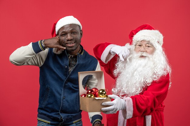 Vorderansicht des Weihnachtsmannes mit dem jungen Mann, der Weihnachtsbaumspielzeug an der roten Wand hält