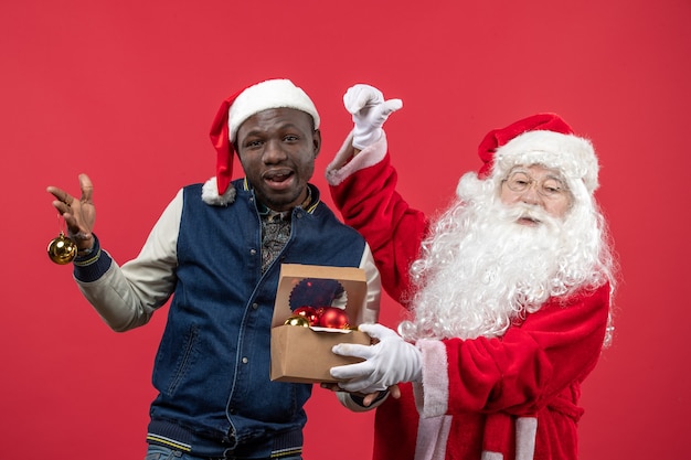 Vorderansicht des Weihnachtsmannes mit dem jungen Mann, der Weihnachtsbaumspielzeug an der roten Wand hält