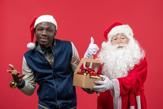 Vorderansicht des Weihnachtsmannes mit dem jungen Mann, der Weihnachtsbaumspielzeug an der roten Wand hält