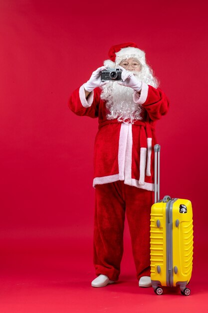 Vorderansicht des Weihnachtsmannes mit gelber Tasche, die Foto mit Kamera an der roten Wand nimmt