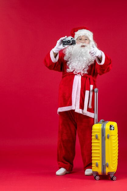 Vorderansicht des Weihnachtsmannes mit gelber Tasche, die Foto mit Kamera an der roten Wand nimmt