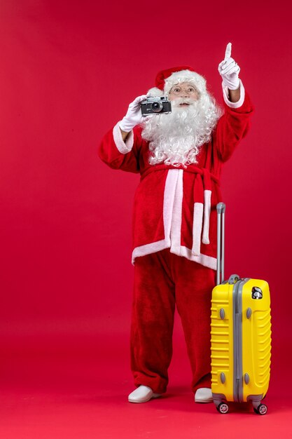 Vorderansicht des Weihnachtsmannes mit gelber Tasche, die Foto mit Kamera an der roten Wand nimmt