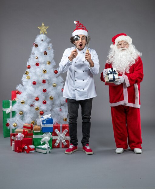 Vorderansicht des Weihnachtsmannes mit männlichem Koch um Weihnachtsgeschenke auf grauer Wand