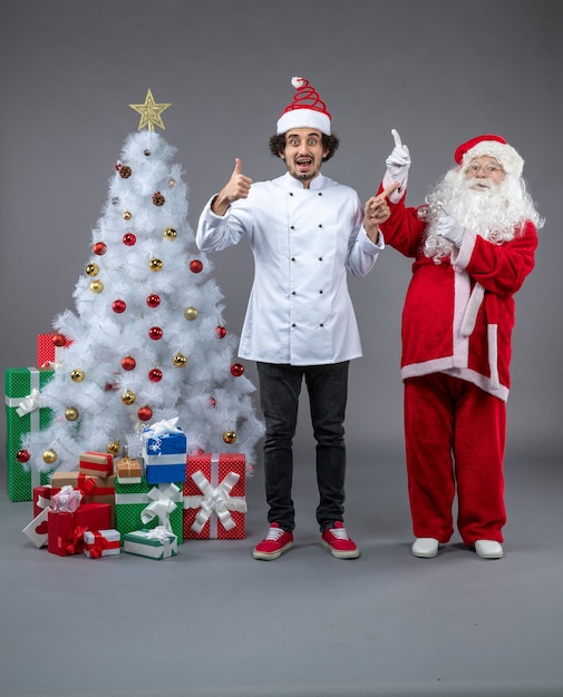 Vorderansicht des Weihnachtsmannes mit männlichem Koch um Weihnachtsgeschenke auf grauer Wand