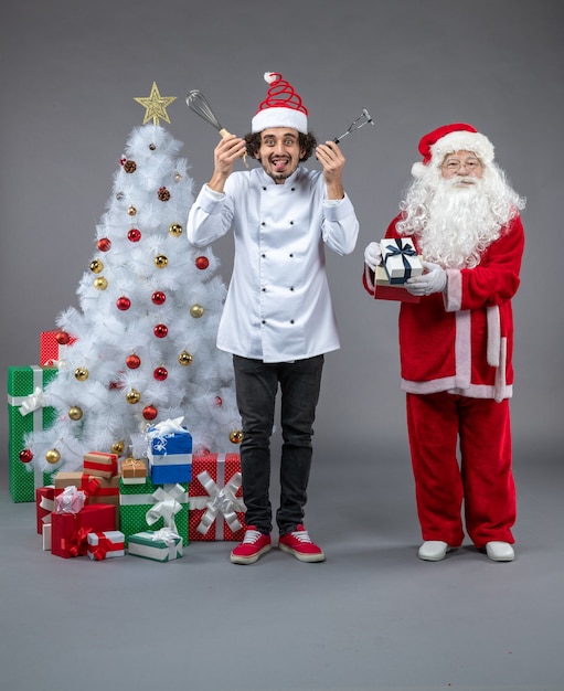 Kostenloses Foto vorderansicht des weihnachtsmannes mit männlichem koch um weihnachtsgeschenke auf grauer wand