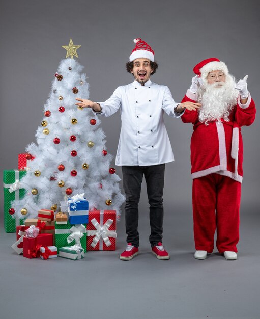 Vorderansicht des Weihnachtsmannes mit männlichem Koch um Weihnachtsgeschenke auf grauer Wand