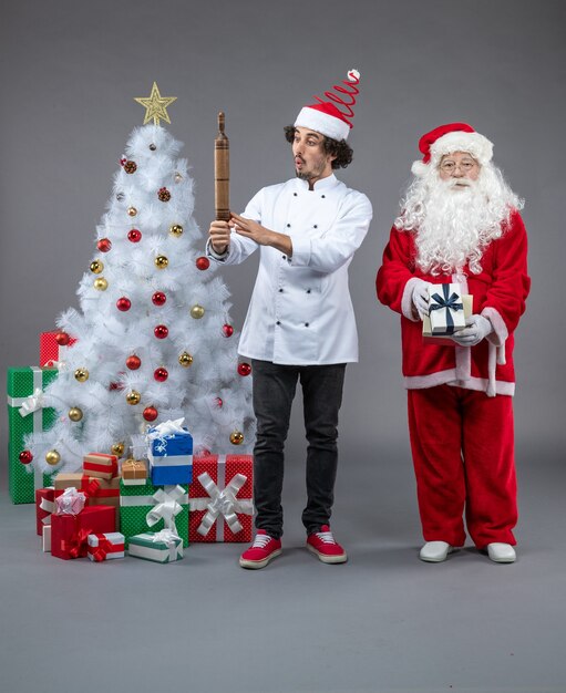 Vorderansicht des Weihnachtsmannes mit männlichem Koch um Weihnachtsgeschenke auf grauer Wand