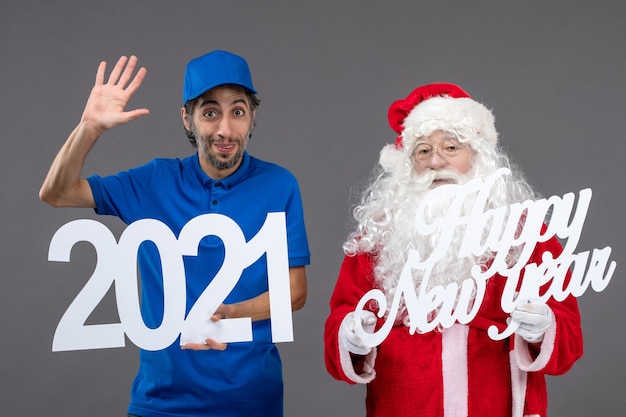 Vorderansicht des Weihnachtsmannes mit männlichem Kurier, der frohes neues Jahr und 2021 Bretter an der grauen Wand hält