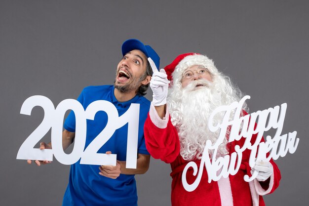 Vorderansicht des Weihnachtsmannes mit männlichem Kurier, der frohes neues Jahr und 2021 Bretter an der grauen Wand hält