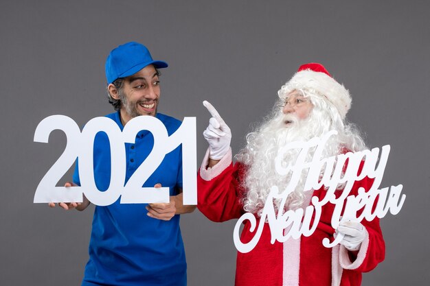 Vorderansicht des Weihnachtsmannes mit männlichem Kurier, der frohes neues Jahr und 2021 Bretter an der grauen Wand hält