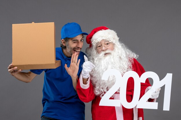 Vorderansicht des Weihnachtsmannes mit männlichem Kurier, der Nahrungsmittelbox und an der grauen Wand hält