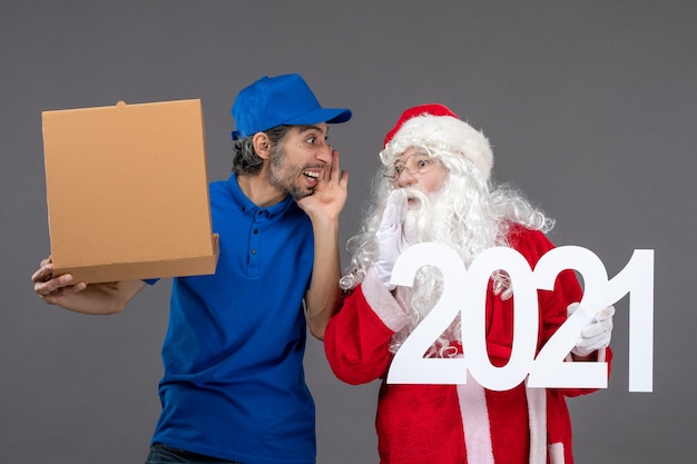 Vorderansicht des Weihnachtsmannes mit männlichem Kurier, der Nahrungsmittelbox und an der grauen Wand hält