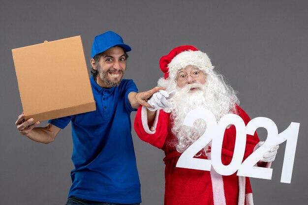 Vorderansicht des Weihnachtsmannes mit männlichem Kurier, der Nahrungsmittelbox und an der grauen Wand hält