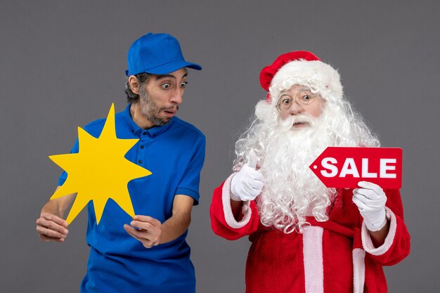 Vorderansicht des Weihnachtsmannes mit männlichem Kurier, der Verkaufsfahne und gelbes Zeichen an der grauen Wand hält