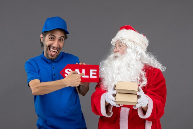 Vorderansicht des Weihnachtsmannes mit männlichem Kurier, der Verkaufsfahne und Lebensmittelverpackungen auf grauer Wand hält