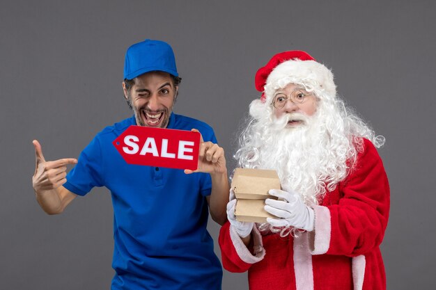 Vorderansicht des Weihnachtsmannes mit männlichem Kurier, der Verkaufsfahne und Lebensmittelverpackungen auf grauer Wand hält