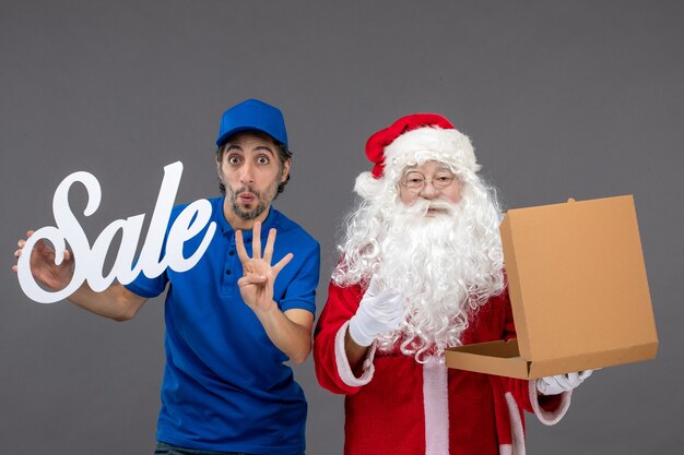 Vorderansicht des Weihnachtsmannes mit männlichem Kurier, der Verkaufsschreiben und Nahrungsmittelbox auf grauer Wand hält