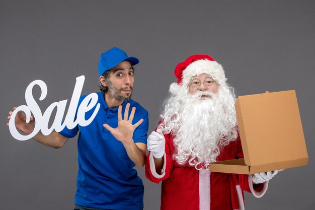 Vorderansicht des Weihnachtsmannes mit männlichem Kurier, der Verkaufsschreiben und Nahrungsmittelbox auf grauer Wand hält