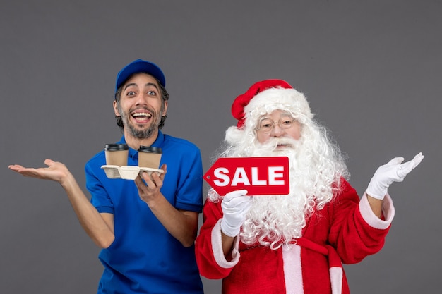 Vorderansicht des Weihnachtsmannes mit männlichem Kurier, der Verkaufsschrift und Kaffee an der grauen Wand hält