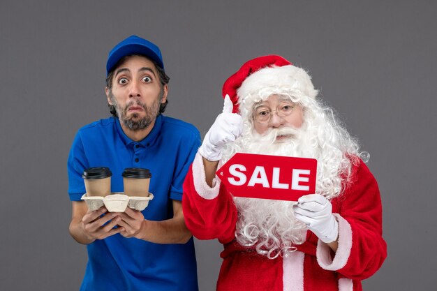 Vorderansicht des Weihnachtsmannes mit männlichem Kurier, der Verkaufsschrift und Kaffee an der grauen Wand hält